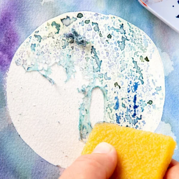 A close up of a persons hand erasing some adhesive from a painted piece of watercolour paper using the Mont Marte Signature Adhesive Eraser. You can see the adhesive being removed. It is an image of a moon. The image is cut off by the frame.