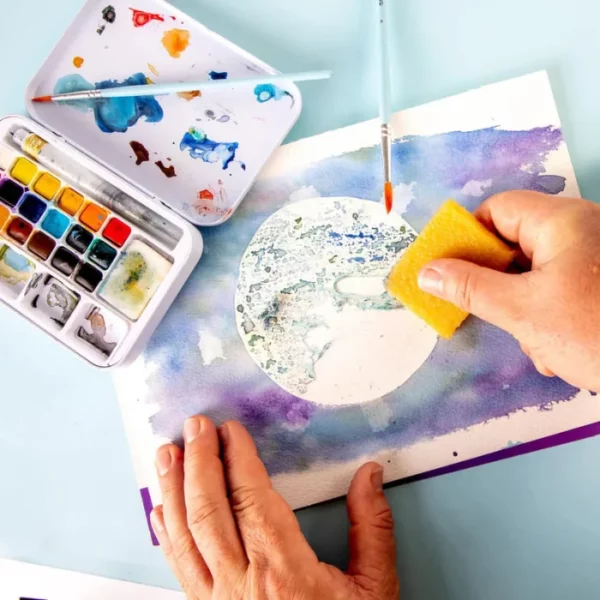 A close up of a persons hand erasing some adhesive from a painted piece of watercolour paper using the Mont Marte Signature Adhesive Eraser. You can see the adhesive being removed. It is an image of a moon. The image is cut off by the frame.