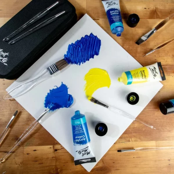 Brushes from the Mont Marte Signature Clear Handle Acrylic Brush Set are shown loose in the frame, scattered over some watercolour paper that has been painted with blue and yellow. There are some tubes of paint scattered around the frame. The image is cut off by the frame.