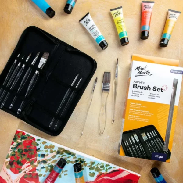 Brushes from the Mont Marte Signature Clear Handle Acrylic Brush Set are shown loose in the frame, scattered over some watercolour paper that has been painted with blue and yellow. There are some tubes of paint scattered around the frame. The image is cut off by the frame.