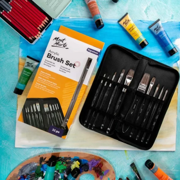 Brushes from the Mont Marte Signature Clear Handle Acrylic Brush Set are shown loose in the frame, scattered over some watercolour paper that has been painted with blue and yellow. There are some tubes of paint scattered around the frame. The image is cut off by the frame.