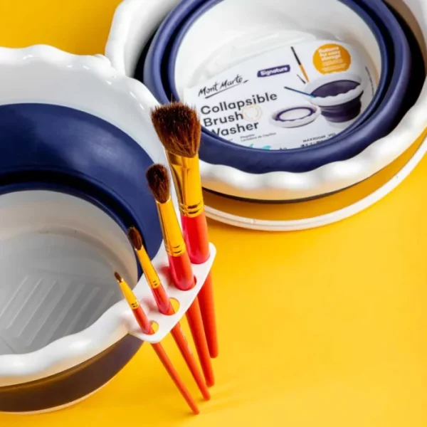 A close up of a Mont Marte Signature Collapsible Brush Washer on a yellow background. There is an open washer at the front, left hand side of the frame and there are 4 paint brushes along the lip of the washer in the slots provided by the washer. A collapsed washer is seen in the background.