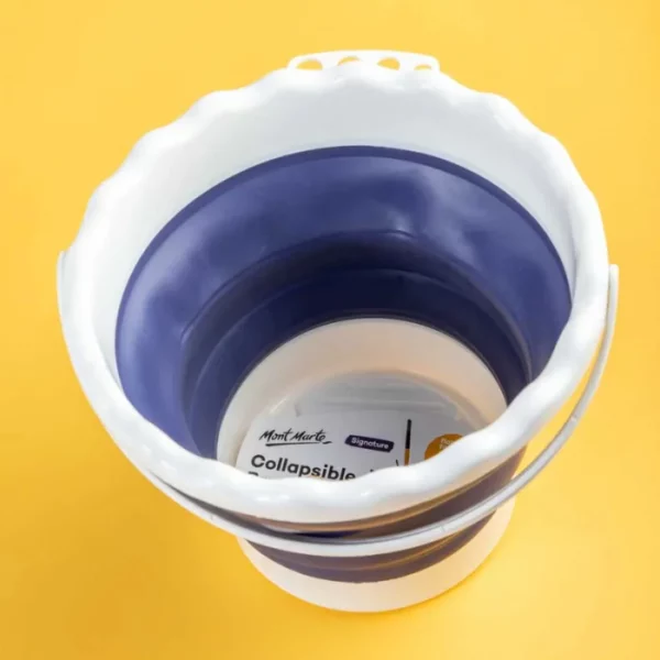 A Mont Marte Signature Collapsible Brush Washer is shown in the center of the frame on a yellow background. The washer is made from a white and blue plastic and resembles a bucket with a scalloped lip.