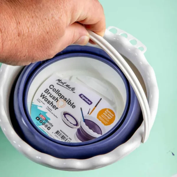 A close up of a persons hand holding the handle of a Mont Marte Signature Collapsible Brush Washer. The washer is made of plastic and is in it's collapsed state in the frame. On a teal background.