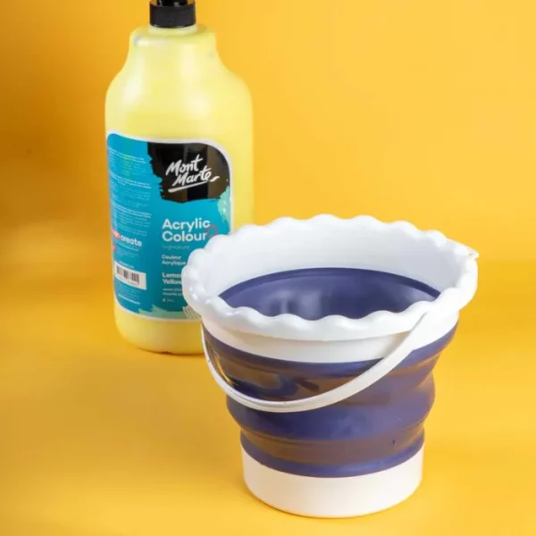 A Mont Marte Signature Collapsible Brush Washer is shown at the front of the frame and a bottle of acrylic pouring medium is in the background and slightly blurred. The washer is white and blue plastic. On a yellow background.