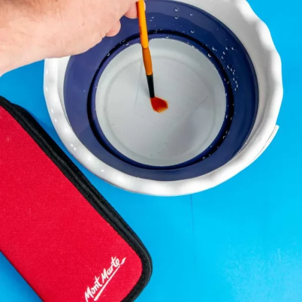 A Mont Marte Signature Collapsible Brush Washer is shown at the top of the frame with a persons hand coming into the frame dipping a paint brush in the washer. there is a red paint brush case below the washer. On a blue background.