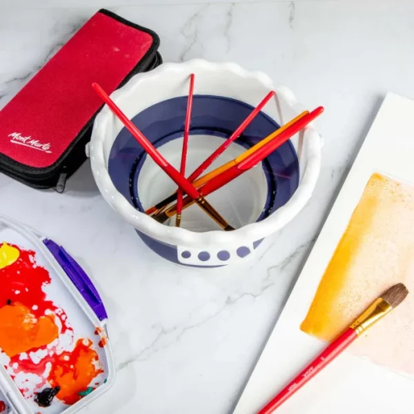 A Mont Marte Signature Collapsible Brush Washer is shown in the center of the frame with 4 red handled paint brushes inside the washer. The washer is made form a white and blue plastic and has a scalloped lip.