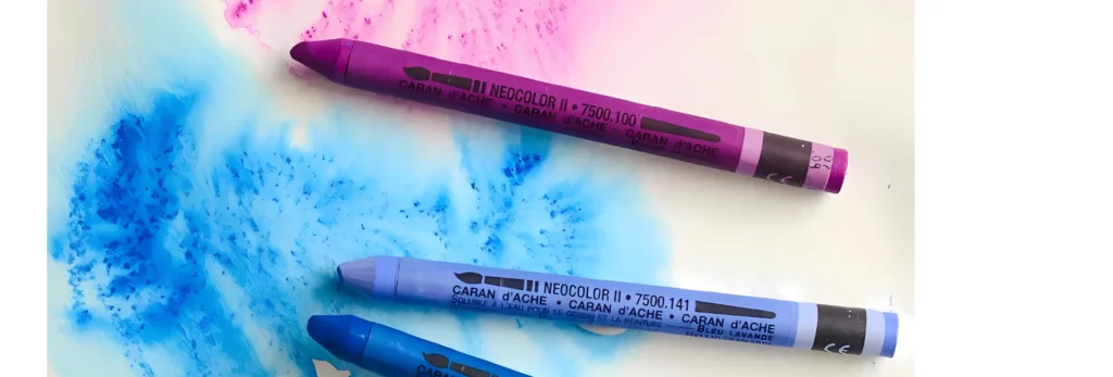 the image is a close up of three pastels that are lying on a paper and have coloured in on the paper and water been used on it to make it look like a watercolour design. they are in shade of blue and purple.