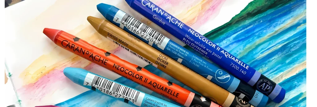 the image is a close up of five neocolour 2 crayons. they are lying on a paper that has been coloured in on. the paper has the image of an ocean and mountains on it. the colours of the pastels are blue, orange, brown, and two different shades of blue.