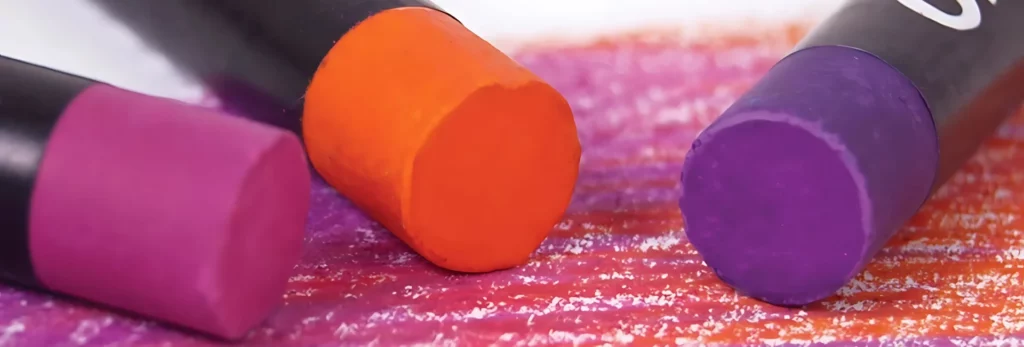 the image is a close up of three oil pastels sitting on top of an area that they have coloured in on. the colours are pink, red and purple. just the tip of the pastel is visable in the image.