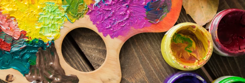 the image is a close up of a wooden paint palette that has paint all over it and there are pots of paint around it on a wooden table surface