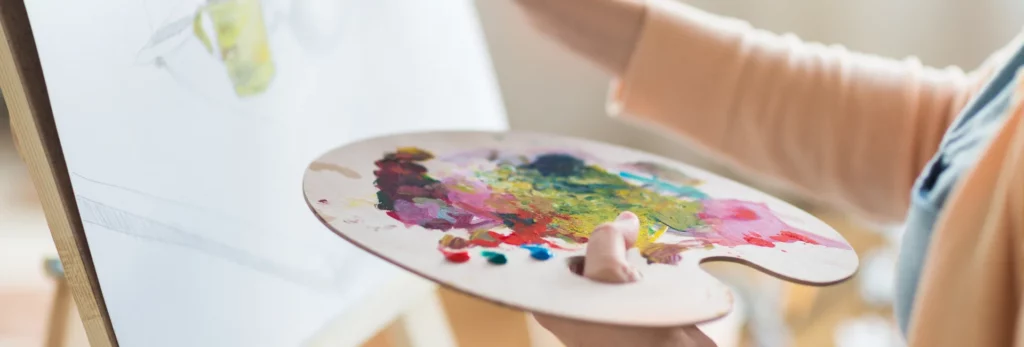 the image is a close up of someone doing a painting in canvas resting on an easel. they are holding a wooden paint palette in their left hand that has blobs of paint all over it, they are using their other hand to paint on the canvas with a paintbrush