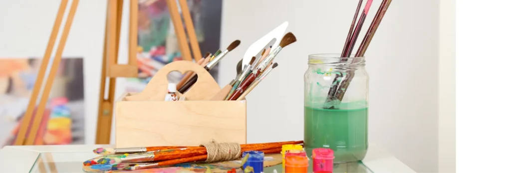 the image is a close up of a table with various art supplies on it. there is a container holding brushes and a glass jar with water in it. a few paints are opened and in front of it. the background is a blurred visual of the room behind