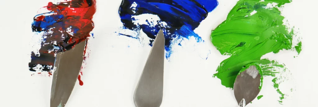 a close up of three palette knives in different sizes and shapes. the left one is mixing red paint, the middle is mixing blue paint and the right hand side is mixing green paint. on a white surface..