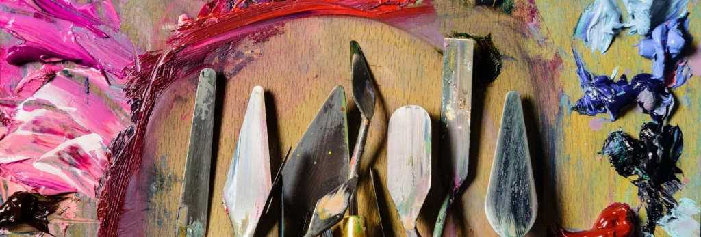 the image is a close up of a variety of palette knives that have been used to mix and apply paint as there is paint on them. they are on a wooden palette and there are various colours of paint that have been mixed and applied around the palette. in shades of pink, white and red.