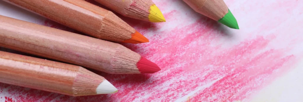 the image is of a close up of five pastel pencils. the tops are only visable, the colours are white, pink, orange, yellow and green. they are all pointing to the right of the image. they are sitting on a paper that has the colours mentioned laid down on it.