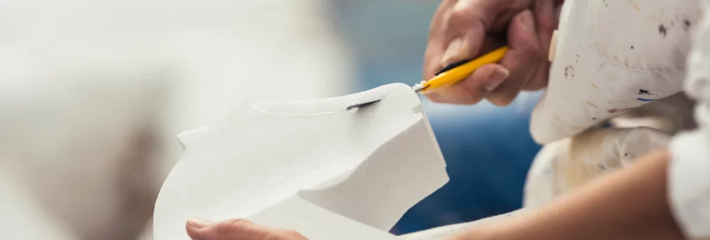 the image shows a man holding a knife and cutting the shape of a horse out of polystyrene. the knife is yellow and the polysytene is white. the background is a blurred view of the room he is working in