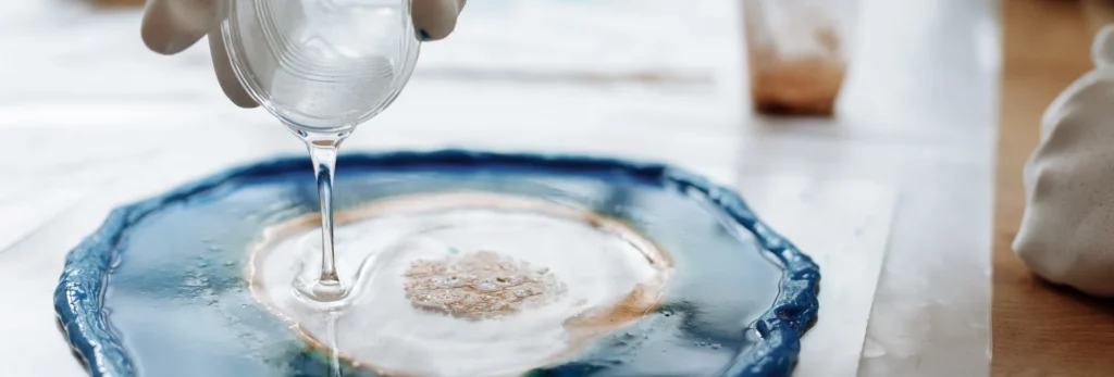 the image is a close up of someone pouring resin on a surface, the resin on the surface looks almost like a rock or preciuos stone. the colours are blue, white and brown. the background is a blurred visual of the room behind them