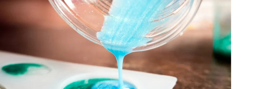 the image is a close up of resin mixed with a sparkly blue colour being poured into moulds on a table. the mould are round in shape. the background is a blur of the room behind