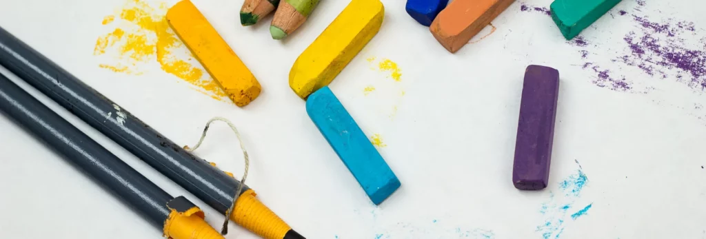 the image shows a birds eye view of a sheet of paper with various tyoes of pastels and pencils and charcoal pencils and stick laid on it. there are areas where the colour has been laid down on the paper.