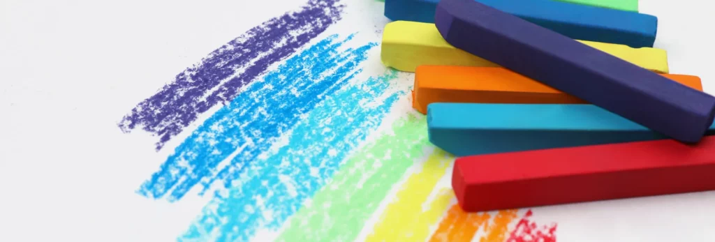 the image shows a close up of pastels on the right hand side. they are stacked on eachother and are bright colours. next to them on the left are areas where they have been drawn on paper in the colours of sticks that are stacked. in a bright rainbow