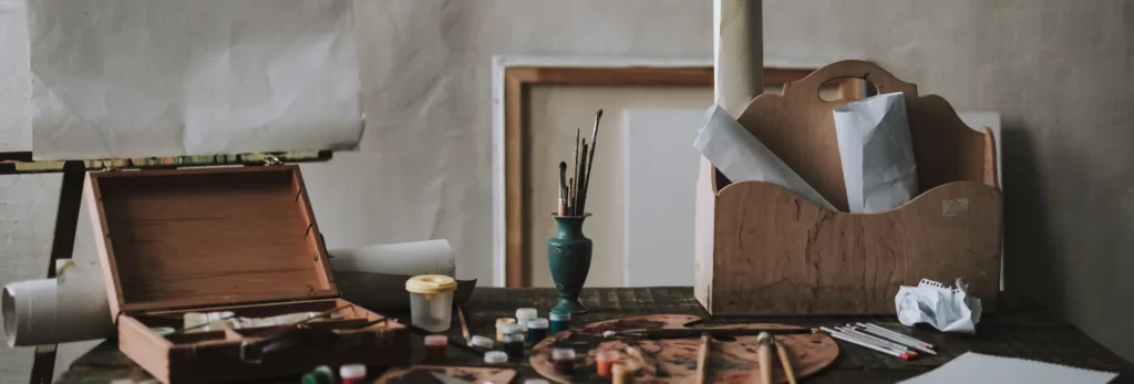 the image is of a table in an art room and it has storage cases on it and palettes infront with paint and paintbrushes around it. the background you can see canvas leaning agaisnt a wall and art papers on easels