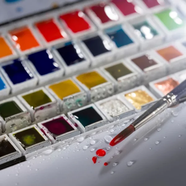 A close up of a paint brush that has been dipped into a red watercolour half pan and has red paint on the brush, with water droplets around the brush. The watercolour set is open and you can see the different coloured half pans. The image is cut off by the frame.