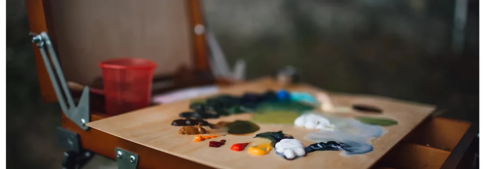 in the center of the image is a close up of a standing easel that has a palette on it that has blibs of paint all the way around it. the palettte and easel are both wooden. there is a small cup in the background with some turpintine in it. the background is a blur of the room