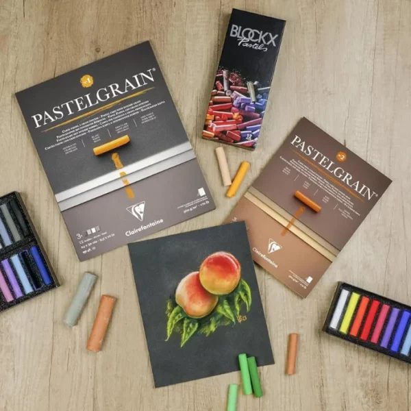 A promotional image for Clairefontaine PastelGrain Pads. There are two pads laying on a wooden desk with various pastels laying around them. There is a drawing of two peaches on a piece of the Pastelgrain paper. Birds eye view.