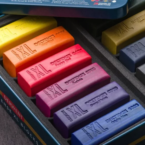 A close up of an open Set of 12 Derwent Inktense XL Blocks. You can see the lid leaning against the open tin in the top of the frame. There are coloured blocks lined up next to each other in the inside tray. Each block is a different colour and they are debossed with the XL brand and colour details. The image is cut off by the frame.