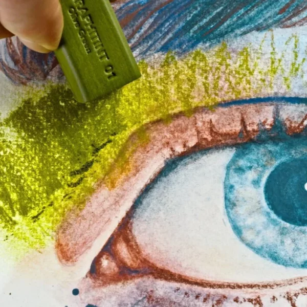 The tips of a persons fingers are seen coming out of the top left hand corner of the frame. They are holding a green graphite stick from the Set of 6 Derwent Graphitint XL Blocks. They are colouring in a picture of a persons eye. The image is a close up and is cut off by the frame.