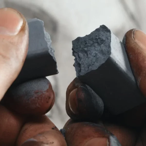 A close up of a persons hands breaking a piece of XL charcoal from the Set of 6 Derwent Charcoal XL Blocks. The image is cut off by the frame. Ther persons hands are full of charcoal and there is a blurred image of a drawing behind the hands.